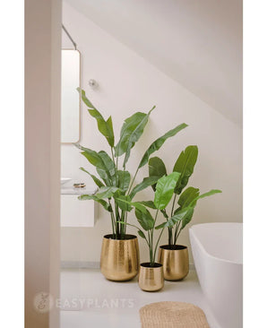 Une plante artificielle Strelitzia Nicolai Deluxe de 120 cm se dresse dans une jardinière dorée brillante sur le sol d'une salle de bain moderne et minimaliste avec une baignoire blanche et des murs de couleur claire.