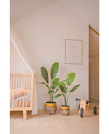 Une chambre d'enfant cosy avec un berceau en bois, une luxueuse plante artificielle Strelitzia Nicolai 120 cm, un dessin au trait minimaliste encadré au mur et une petite draisienne en bois avec le label MIA sur un parquet en bois clair.