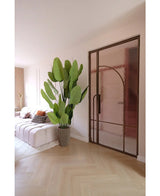 Un salon moderne avec un canapé blanc, des coussins à motifs et la Plante Artificielle Strelitzia Real Touch Deluxe 210 cm. Un parquet en bois clair, des murs rose tendre et une porte vitrée avec un cadre en métal bronze créent une atmosphère lumineuse et aérée.