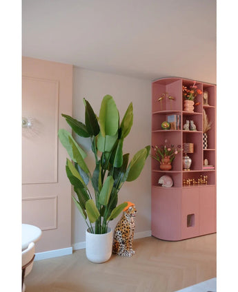 La plante artificielle Strelitzia Real Touch Deluxe 210 cm se trouve à côté d'une figurine de guépard en céramique avec un chapeau orange, à côté d'une haute étagère rose remplie de vases, de fleurs et d'ornements dans une pièce aux couleurs pastel.