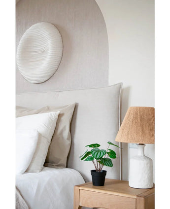 Une chambre confortable comprend un lit avec une literie neutre, une table de chevet en bois clair avec une plante de pastèque artificielle de 29 cm et une lampe blanche avec un abat-jour en osier. Une sculpture murale texturée blanche est suspendue au-dessus du lit.