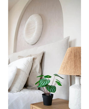 Un lit soigneusement fait avec des oreillers et une literie blancs se trouve à côté d'une table de chevet en bois avec une plante de pastèque artificielle de 29 cm et une lampe avec un abat-jour tissé ; au-dessus de la tête de lit est suspendue une sculpture murale texturée blanche.