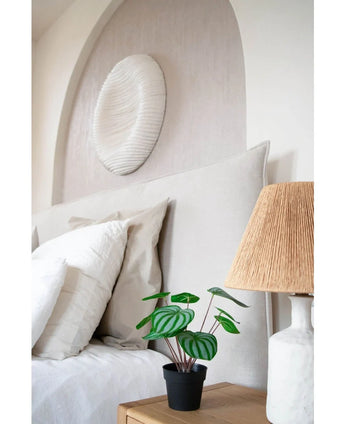 Un lit soigneusement fait avec des oreillers et une literie blancs se trouve à côté d'une table de chevet en bois avec une plante de pastèque artificielle de 29 cm et une lampe avec un abat-jour tissé ; au-dessus de la tête de lit est suspendue une sculpture murale texturée blanche.