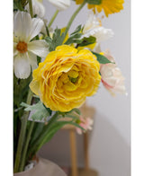 Een close-up van de Kunstranonkel Real Touch Geel 60cm, een levendige gele kunstranonkel, omringd door witte bloemen en groene bladeren in een vaas binnenshuis, met een zacht onscherpe achtergrond.