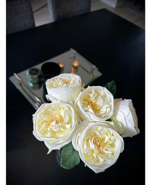 De Kunstrozenboeket "Carmen" Real Touch Crème wit 36cm met groene bladeren staat op een donkere tafel. Op de achtergrond zijn een spiegelplateau met kaarsen en twee grijze stoelen zichtbaar.