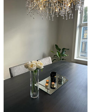 Een moderne eettafel met de Kunstrozenboeket "Carmen" Real Touch Crème wit 36cm als middelpunt, een spiegelplateau met kaarsen en decor, een grijs gestoffeerde stoel, een bladplant bij het raam en een kristallen kroonluchter boven.
