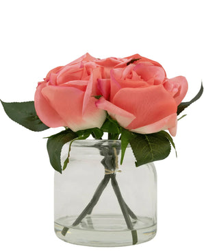 De Kunstrozenboeket Real Touch Roze 18cm heeft vier roze kunstrozen met groene bladeren, gerangschikt in een kleine glazen pot en tegen een witte achtergrond.
