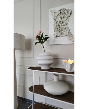 Un intérieur moderne avec une décoration murale texturée blanche, un vase sculptural blanc avec une branche d'eucalyptus artificielle à fleurs 62 cm vieux rose, une bougie décorative et un vase rond en céramique sur une étagère en bois à côté d'un lampadaire.