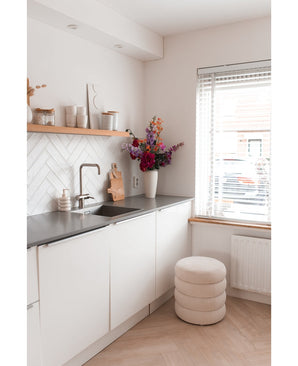 Une cuisine moderne et minimaliste avec des armoires blanches, un plan de travail noir et une fleur d'hortensia artificielle de 55 cm rose foncé dans un vase près du plan de travail, ainsi que des étagères en bois décorées et un tabouret rond avec des coussins près d'une fenêtre avec stores.