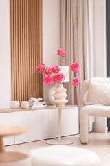 Un salon moderne dispose d'une table d'appoint blanche avec la branche artificielle de fleur de cerisier de 85 cm rose, à laquelle ont été ajoutées des fleurs rose vif. Une décoration neutre, des accents de bois clair, des rideaux beiges et un fauteuil structuré complètent l'espace cosy et minimaliste.