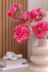 Un vase en céramique blanche contient une branche de fleur de cerisier artificielle de 85 cm rose. Le fond montre des panneaux de bois verticaux, une pile de livres et un petit objet décoratif au-dessus.