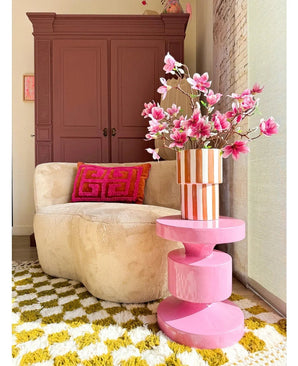 Een gezellige woonkamer met een beige ronde bank, een roze en oranje geometrisch kussen, een roze bijzettafel, Kunsttak Magnolia kleinbloemig 89 cm roze in een vaas, een geel-wit gedessineerd vloerkleed en een kast in kastanjebruin op de achtergrond.