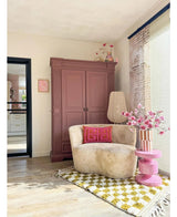 Een gezellige leeshoek heeft een crèmekleurige stoel, roze kussens, een geel gedessineerd vloerkleed en een mauve kast. Op een roze bijzettafel staat de Kunsttak Magnolia kleinbloemig 89 cm roze, terwijl grote ramen deze uitnodigende ruimte vullen met zacht licht.