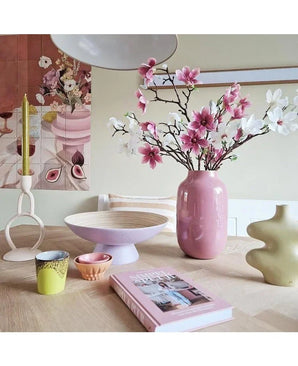 Sur une table en bois clair se trouve le Art Branch Magnolia à petites fleurs blanches de 89 cm dans un vase rose, des céramiques modernes, des tasses colorées, un bol, une bougie jaune dans un support unique et un livre rose, avec une œuvre florale sur le mur derrière.