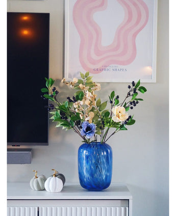 Un vase en verre bleu avec une fleur artificielle violette de 76 cm de branche artificielle se dresse sur une armoire blanche. À côté se trouvent deux citrouilles blanches aux tiges dorées. Sur le mur derrière, il y a une télévision et une impression d'art abstraite rose.