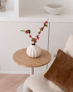 La Branche Artificielle Baies 64 cm rose - un vase blanc rempli de branches de baies artificielles roses et de feuilles vertes - se dresse sur une petite table ronde à côté d'une chaise beige avec un coussin marron dans une pièce cosy et minimaliste.