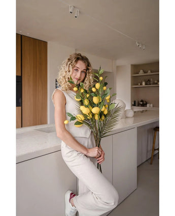 Une femme aux cheveux blonds bouclés, vêtue d'une tenue couleur crème, s'appuie contre un comptoir et tient le bouquet 'Branche Artificielle Citron 104 cm jaune', décoré de verdure luxuriante. La cuisine moderne est décorée dans des couleurs neutres et présente des touches de bois.