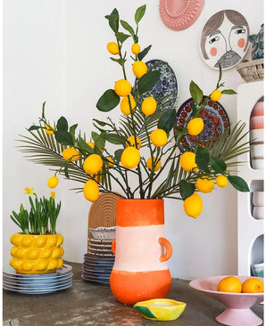 Une pièce lumineuse avec un grand vase rempli de citrons et de feuilles vertes, dans lequel le remarquable Kunsttak Lemon 104 cm jaune occupe une place de choix dans l'agencement, entouré de bols en céramique colorés, d'un bol de citrons et d'assiettes décoratives au mur - créant un look joyeux et éclectique.