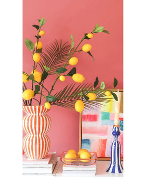 Contre un mur rose corail se trouvent un vase rayé rempli de branches de citron Kunsttak de 104 cm en jaune, un tableau coloré, une pile de livres, un chandelier bleu avec une bougie et un plateau de citrons.