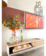 Sur une étagère en bois se trouve un vase coloré contenant l'Art Branch Mandarin 93 cm orange, à côté d'une œuvre d'art encadrée à rayures roses sur laquelle on peut lire « Kissje op je Hart ». Sur l'étagère se trouvent également une bougie verte et des couvertures pliées.