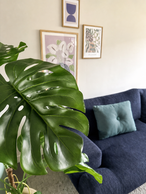Une plante artificielle Monstera de 120 cm est placée bien en évidence à l'avant. Derrière se trouve un canapé bleu foncé avec un coussin bleu clair contre un mur beige avec trois œuvres d'art encadrées.