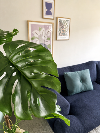 Une plante artificielle Monstera de 120 cm est placée bien en évidence à l'avant. Derrière se trouve un canapé bleu foncé avec un coussin bleu clair contre un mur beige avec trois œuvres d'art encadrées.