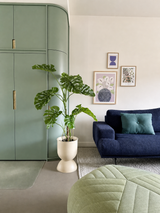 Un salon moderne avec un placard intégré vert, une plante artificielle Monstera 120 cm, un canapé bleu avec un coussin bleu sarcelle, de l'art abstrait au mur et un pouf vert sur un tapis clair.