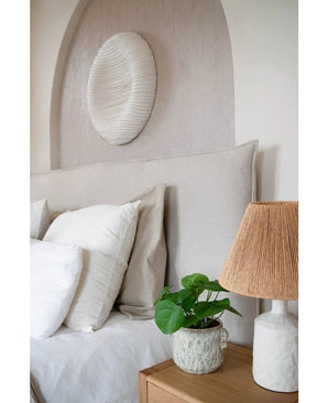 Un coin nuit douillet avec une tête de lit beige, des oreillers blancs et beiges, une table de chevet en bois, une lampe en céramique avec abat-jour tressé, la plante à crêpes artificielle 27 cm et une décoration murale blanche au-dessus du lit.
