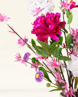 Le bouquet de fleurs artificielles en soie Pink Reverie contient une grande pivoine magenta, de petites fleurs roses, des fleurs violettes ressemblant à des marguerites et des feuilles vertes dans un vase - un bouquet romantique sur un fond clair.