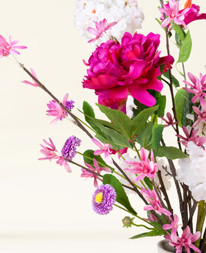 Le bouquet de fleurs artificielles en soie Pink Reverie contient une grande pivoine magenta, de petites fleurs roses, des fleurs violettes ressemblant à des marguerites et des feuilles vertes dans un vase - un bouquet romantique sur un fond clair.