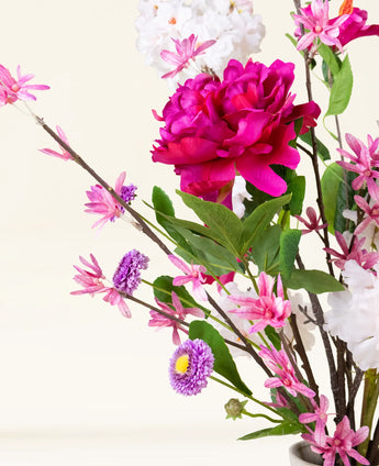 Le bouquet de fleurs artificielles en soie Pink Reverie contient une grande pivoine magenta, de petites fleurs roses, des fleurs violettes ressemblant à des marguerites et des feuilles vertes dans un vase - un bouquet romantique sur un fond clair.