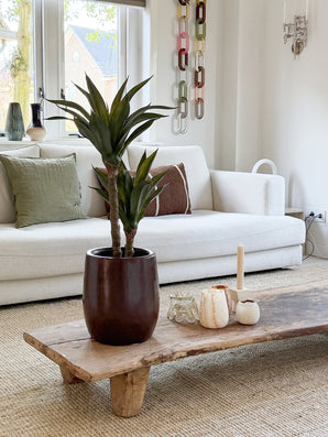 Un salon moderne avec un canapé blanc, des coussins verts et marron, une table basse en bois décorée de bougies et de l'agave artificielle 76 cm, et une décoration murale en maille chaîne. La lumière naturelle pénètre par les fenêtres derrière le canapé.