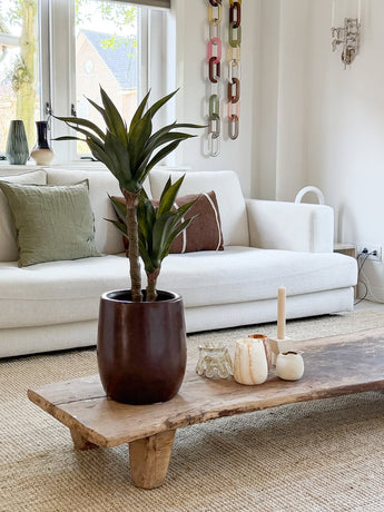 Un salon moderne avec un canapé blanc, des coussins verts et marron, une table basse en bois décorée de bougies et de l'agave artificielle 76 cm, et une décoration murale en maille chaîne. La lumière naturelle pénètre par les fenêtres derrière le canapé.
