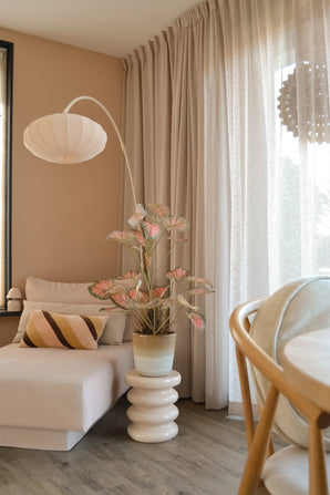 Un coin salon cosy avec une méridienne beige, un coussin rayé, le Caladium Artificiel 90 cm rose qui apporte une touche de vert, une lampe moderne, des rideaux lumineux et un parquet en bois. La douce lumière naturelle pénètre à travers les rideaux transparents.