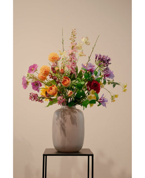 Een beige vaas met Ranonkel kunsttak x5 Real Touch oranje 62cm kunstmatige oranje ranonkels staat op een zwart metalen standaard tegen een effen lichte achtergrond.