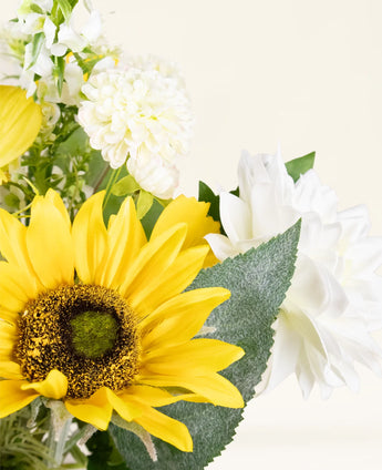 Un gros plan du bouquet de fleurs artificielles en soie Sunny Whisper avec un tournesol jaune vif, des dahlias blancs et des feuilles vertes sur un fond clair et neutre.