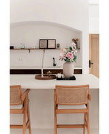 Une cuisine minimaliste dispose d'un îlot blanc avec deux chaises en bois et du Vase Artic Beige Ø21 x H29 cm à fleurs roses et blanches. Des étagères avec des livres et des céramiques sont fixées au mur en arrière-plan.