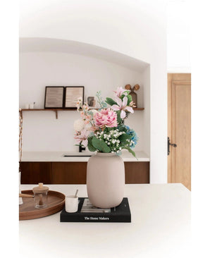 Le Vase Artic Beige Ø21 x H29 cm, rempli de fleurs artificielles roses et bleues, repose sur un livre noir sur un plan de travail blanc. Un éclairage doux accentue la scène minimaliste avec des étagères décoratives et une porte en bois en arrière-plan.