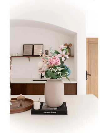 Le Vase Artic Beige Ø21 x H29 cm, rempli de fleurs artificielles roses et bleues, repose sur un livre noir sur un plan de travail blanc. Un éclairage doux accentue la scène minimaliste avec des étagères décoratives et une porte en bois en arrière-plan.
