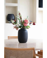 De Vaas Artic Black Ø21 x H29 cm, gevuld met roze, witte en rode bloemen, staat op een ronde beige tafel omringd door twee houten stoelen met gevlochten rieten rugleuningen. Op de achtergrond zijn decoratieve planken zichtbaar.