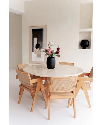 Een ronde marmeren tafel met vier houten stoelen omringt de Vaas Artic Black Ø21 x H29 cm gevuld met bloemen. Abstracte muurkunst en een zwart decorstuk op een ingebouwde plank maken de minimalistische, lichtgetinte kamer compleet.