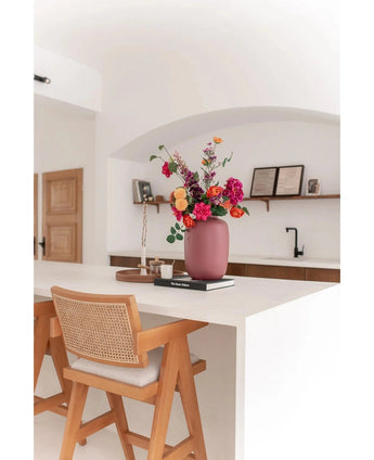 Une cuisine moderne avec un îlot blanc, une chaise en bois et le vase Artic Pink Ø21 x H29 cm rempli de fleurs colorées. Au fond, vous voyez des étagères avec des livres et de la décoration, un plateau et un robinet noir.