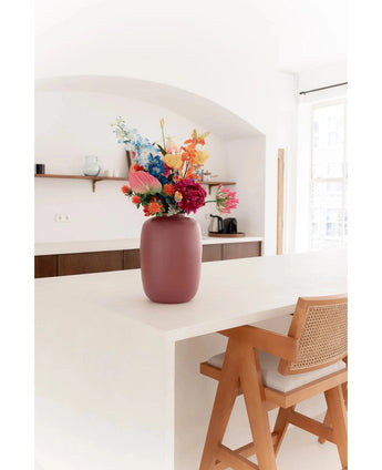 Le vase Artic Pink Ø21 x H29 cm, rempli de fleurs colorées, se dresse sur un îlot de cuisine blanc à côté d'une chaise en bois au dossier tressé. Des étagères minimalistes et une cafetière ornent cet espace lumineux et naturellement éclairé.