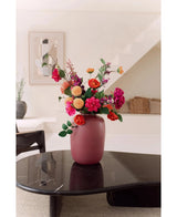Le Vase Artic Pink Ø21 x H29 cm, rempli de fleurs roses, oranges et rouges, se dresse sur une table basse sombre et brillante dans un salon moderne et minimaliste avec un escalier et des œuvres d'art encadrées en arrière-plan.