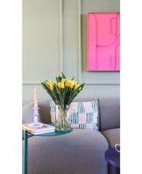 Het Zijden kunstboeket Tulpen in vaas Geel staat op een groen bijzettafeltje naast een roze gedraaide kaars, twee boeken en een grijze gestructureerde bank met een kussen met patroon. Aan de lichtgroene muur hangt een felroze abstract kunstwerk.