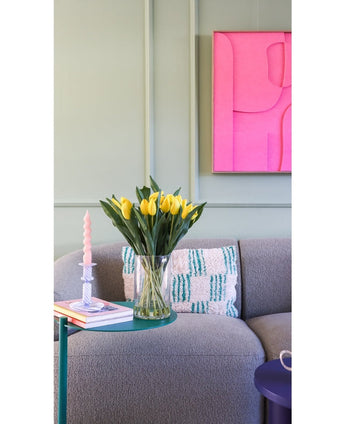 Het Zijden kunstboeket Tulpen in vaas Geel staat op een groen bijzettafeltje naast een roze gedraaide kaars, twee boeken en een grijze gestructureerde bank met een kussen met patroon. Aan de lichtgroene muur hangt een felroze abstract kunstwerk.