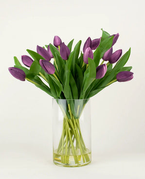 Het Zijden kunstboeket Tulpen in vaas Paars heeft langstelige paarse kunsttulpen met weelderige groene bladeren, prachtig gerangschikt in een heldere glazen vaas tegen een effen witte achtergrond.