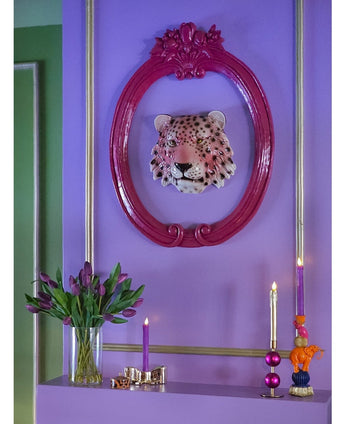 Een levendige paarse muur toont een decoratieve luipaardkop in een roze sierlijke lijst. Hieronder zie je het Zijden kunstboeket Tulpen in vaas Paars, naast een paarse kaars, een gouden kandelaar en kleurrijke kaarsen op een plank.