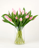 Het Zijden kunstboeket Tulpen in vaas Roze heeft roze tulpen met lange stelen en groene bladeren in een heldere glazen vaas, tegen een effen witte achtergrond.