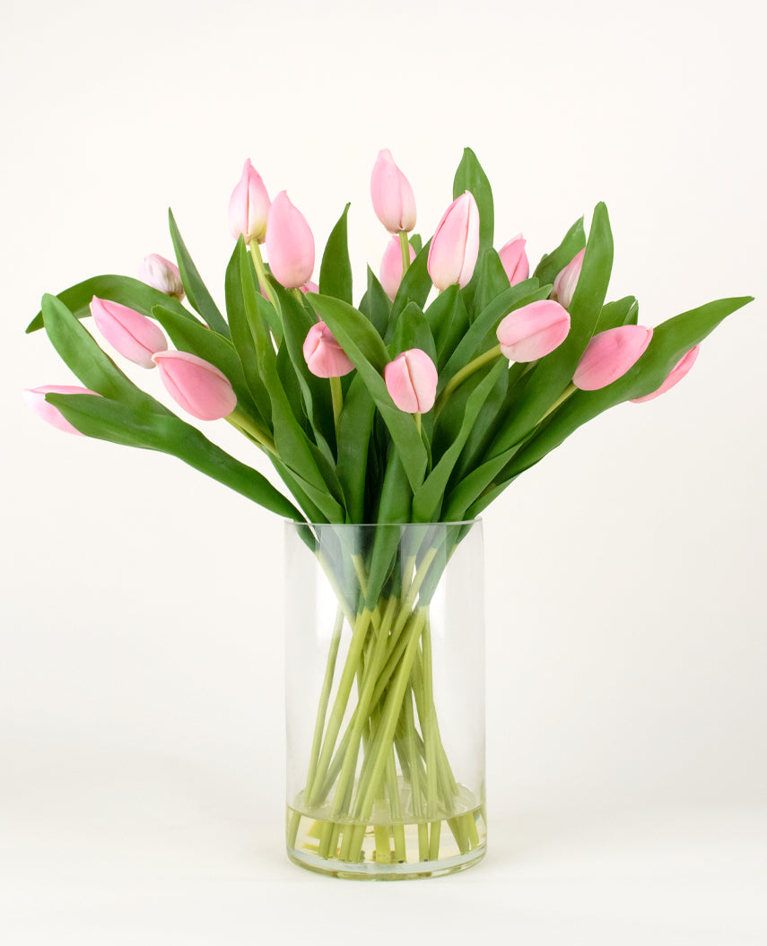 Le bouquet artificiel en soie Tulipes dans un vase Rose présente des tulipes roses avec de longues tiges et des feuilles vertes dans un vase en verre transparent, sur un fond blanc uni.